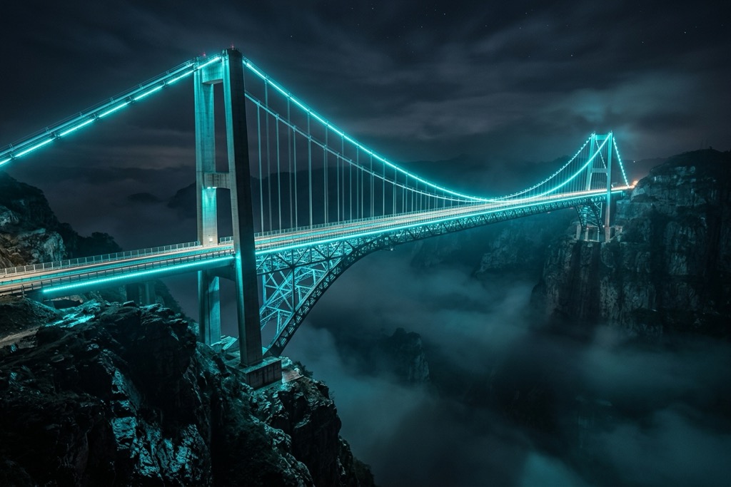 Are Immersive Web Experiences Infrastructure or Luxury? Massive steel and glass suspension bridge at night with cyan LED lighting connecting two dark cliffsides representing the question of whether immersive web experiences are essential infrastructure or optional luxury